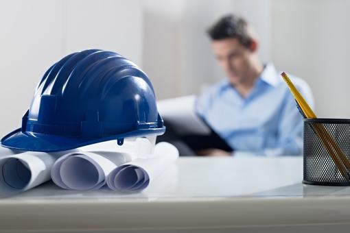 Casco y planos sobre un escritorio, con un ingeniero al fondo